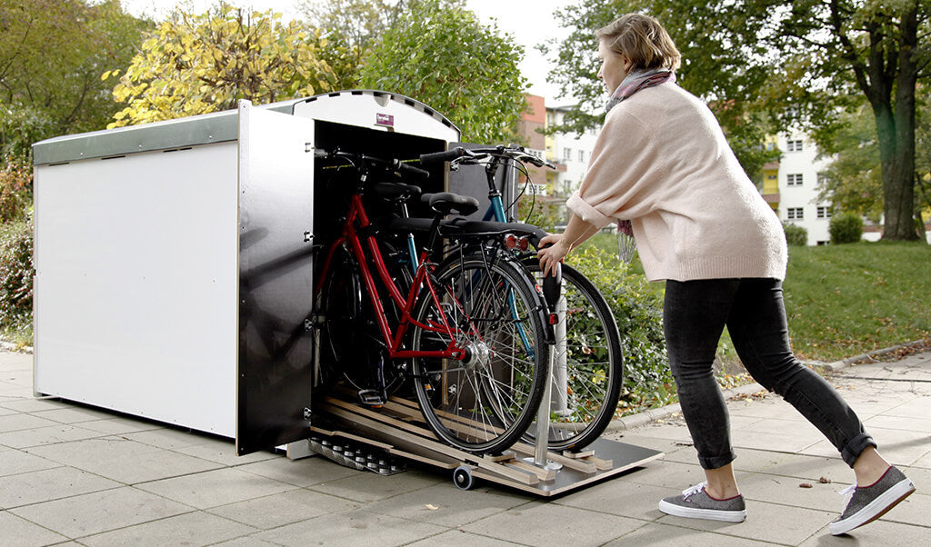 Frau schiebt zwei Fahrräder aus abschließbarem Fahrradgaragenbox auf Gehweg in urbanem Park