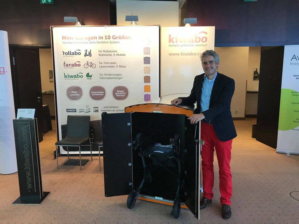 Mann präsentiert faltbare Mini-Garage für Rollator auf Messe mit Kiwabo-Banner im Hintergrund