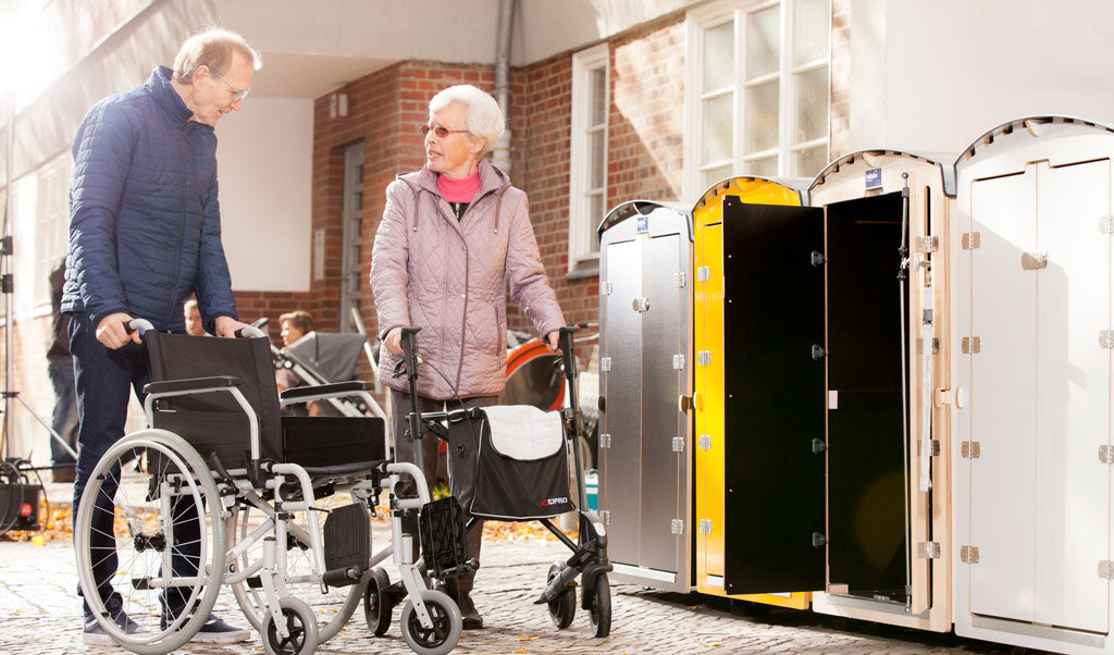 Älteres Paar mit Rollstuhl und Rollator vor geöffneten mobilen Umkleidekabinen im Außenbereich bei sonnigem Wetter