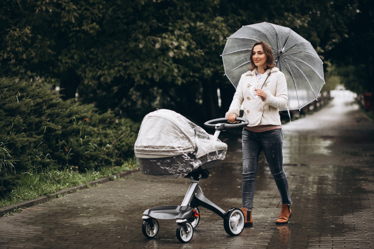 Frau mit transparentem Regenschirm schiebt Kinderwagen mit Regenschutz auf nassem Gehweg