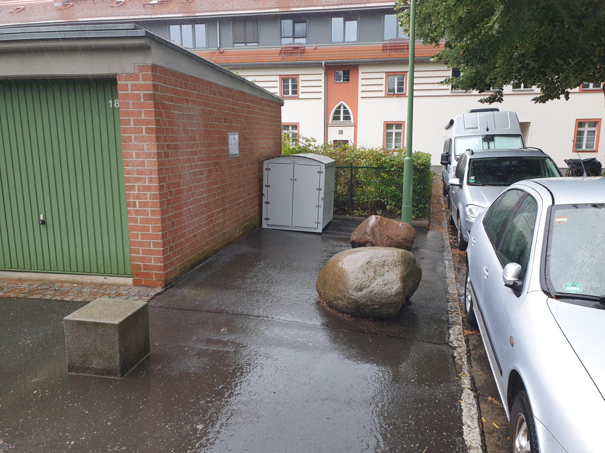 Nasse Parkfläche mit grauer Fahrradgarage, zwei großen Steinen, grünem Garagentor und geparkten Autos