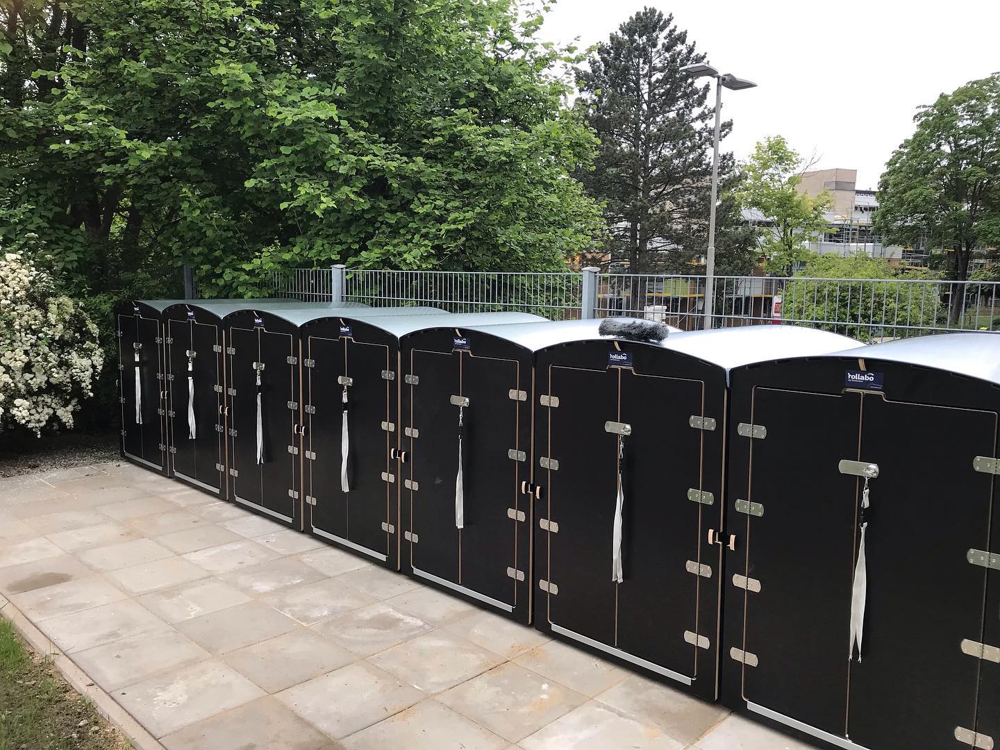 Reihe schwarzer Rollabo Fahrradboxen auf gepflastertem Platz im Freien vor Büschen und Gebäuden