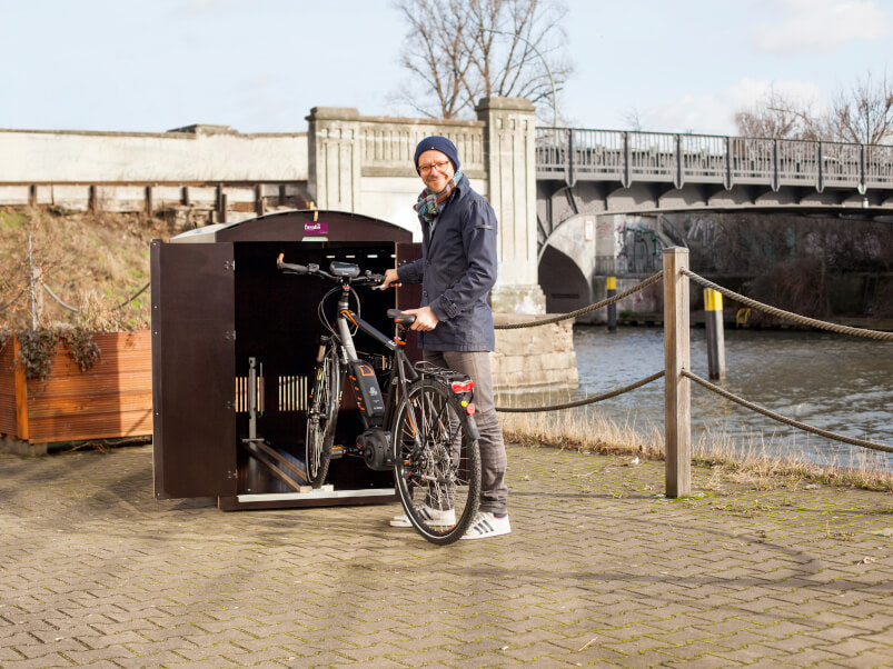 Mann stellt E-Bike in abschließbaren Fahrradbox an Flussufer mit Brücke im Hintergrund