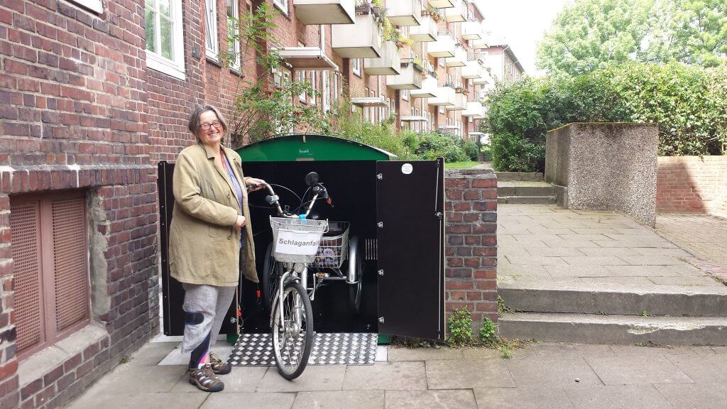 Frau neben Therapie-Dreirad vor offener Garage in Wohngebiet mit rotem Backstein und Grün