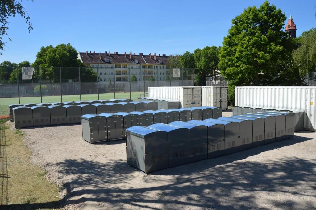 Reihen von grauen Minigaragen auf offenem Gelände vor Wohngebäuden und Bäumen im sonnigen Freiluftbereich