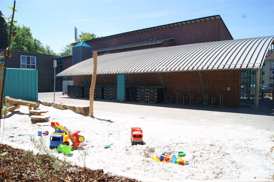 Kinderspielplatz mit Sandkasten und buntem Spielzeug vor moderner Kinderwagenbox und Fahrradständern bei Sonnenschein