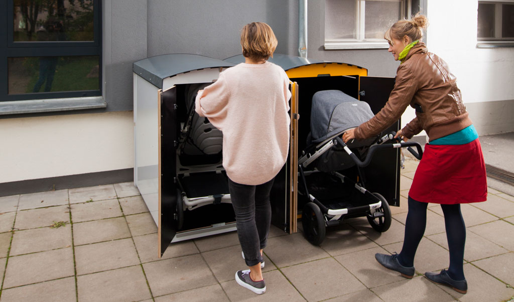 Zwei Frauen verstauen Kinderwagen in abschließbaren Außenboxen vor Wohngebäude auf Pflasterweg