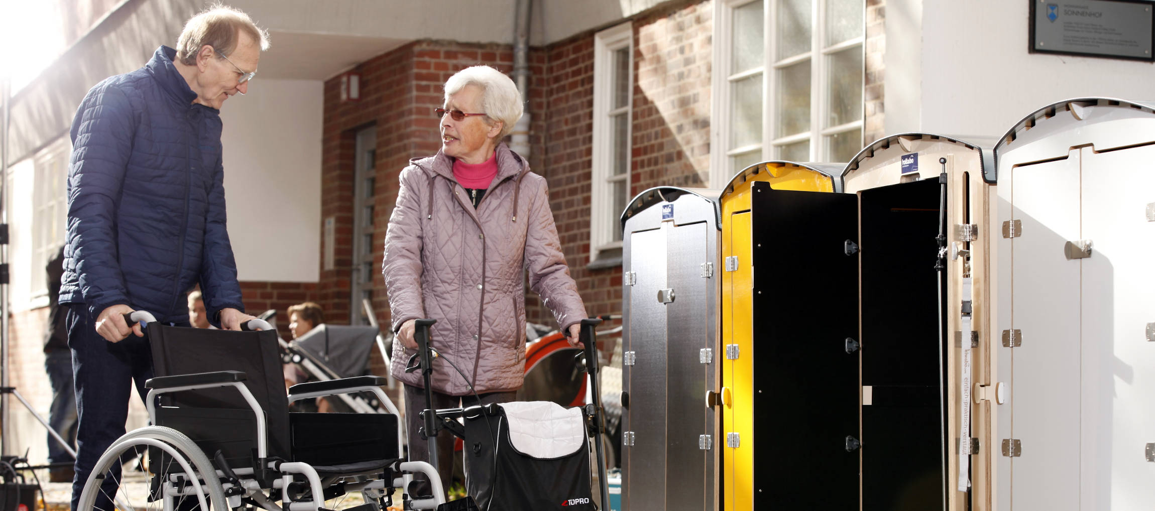 Seniorenpaar mit Rollstuhl und Rollator vor Pflegeschrank im Außenbereich eines Pflegeheims