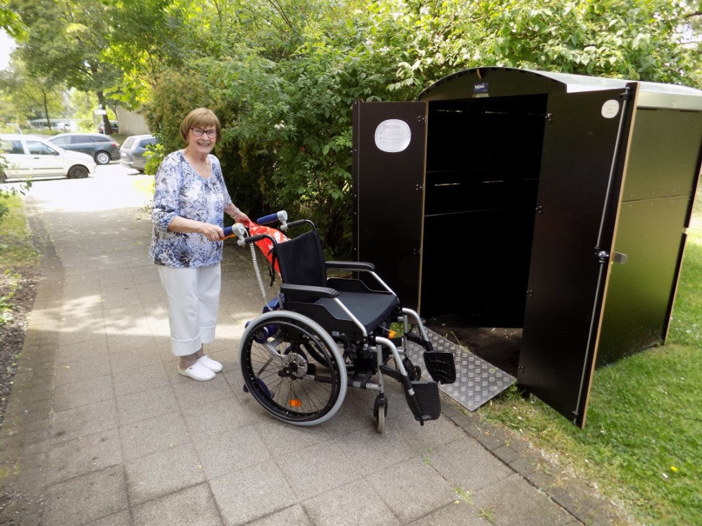 Ältere Frau schiebt Rollstuhl vor offene Rollstuhlgarage im Gartenbereich an Gehweg