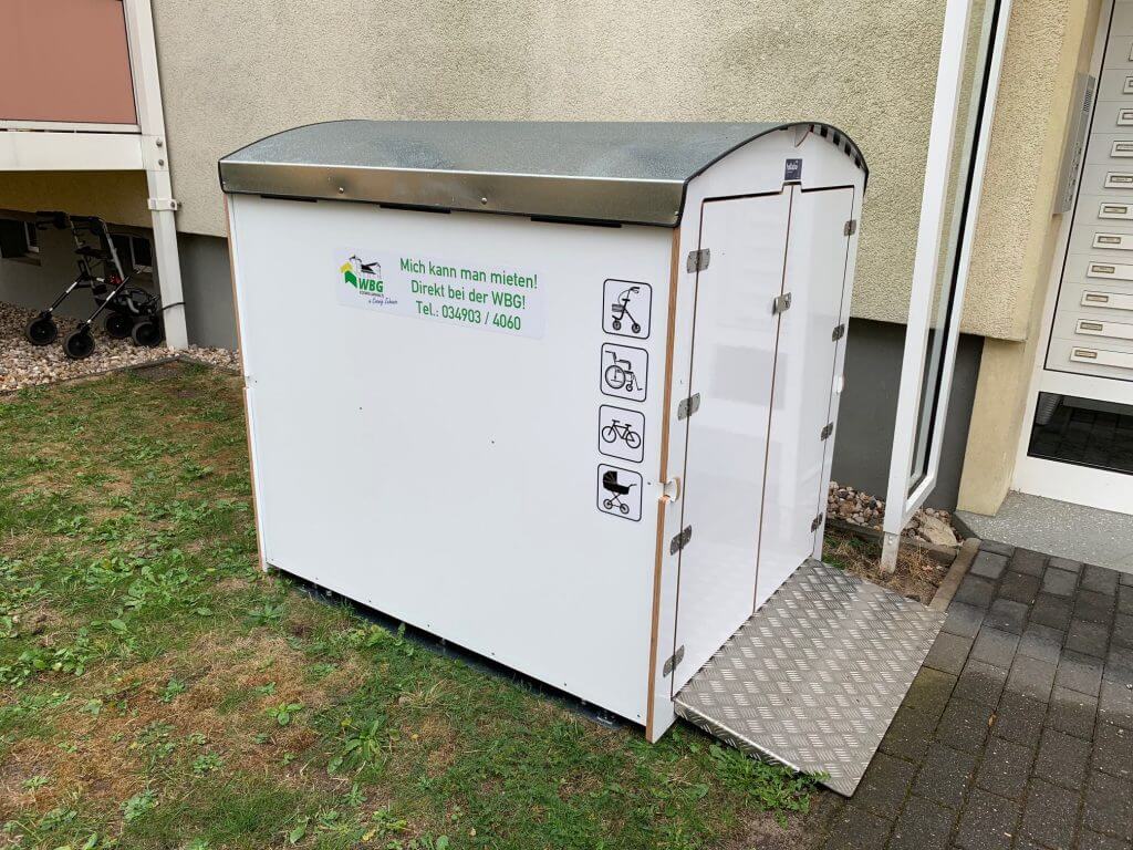 Weiße Minigartenbox mit grauem Metall-Dach und Piktogrammen für Kinderwagen, Rollstuhl und Fahrrad in Wohnumgebung