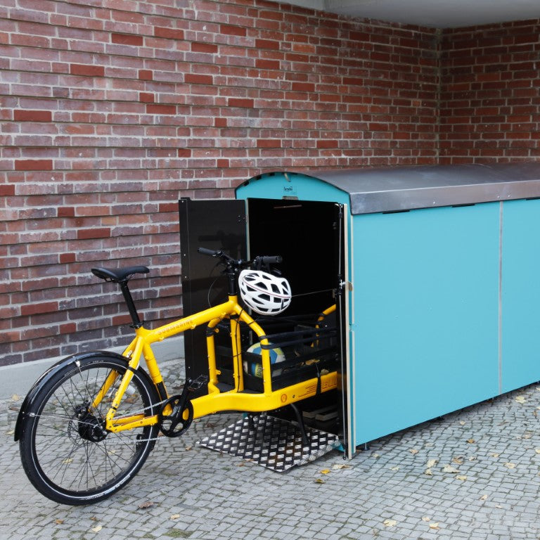 Gelbes Lastenfahrrad mit weißem Helm vor blauem Fahrradgarage in urbanem Umfeld mit Backsteinwand