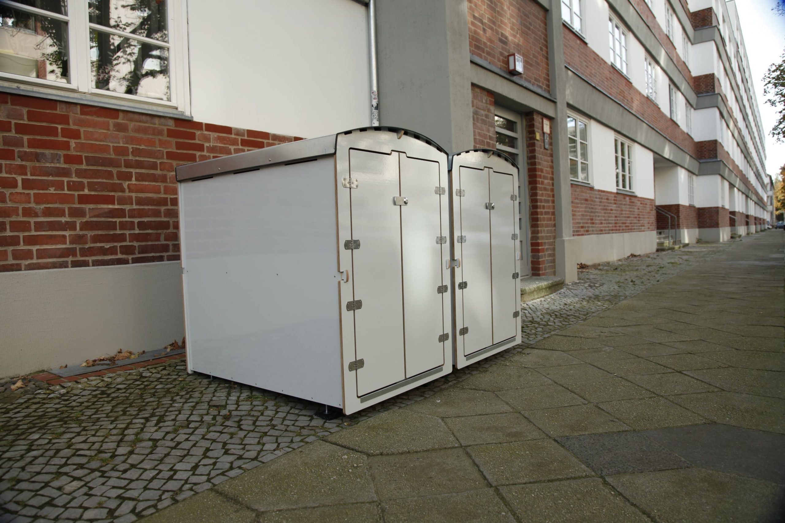 Zwei weiße Aufbewahrungsboxen für Kinderwagen oder Fahrräder vor Wohnhaus auf Gehweg