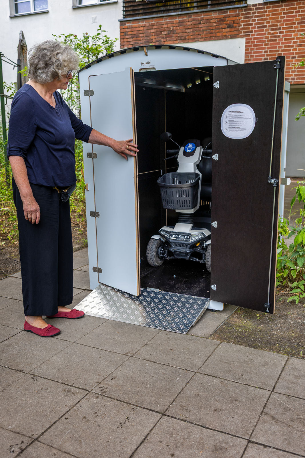 Seniorin mit E-Mobil an einer weißen rollabo Box, Hochformat