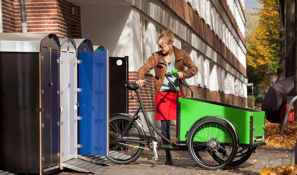 Frau in rotem Rock öffnet grünes Lastenfahrrad neben bunten Schließfächern vor Backsteingebäude im Herbst
