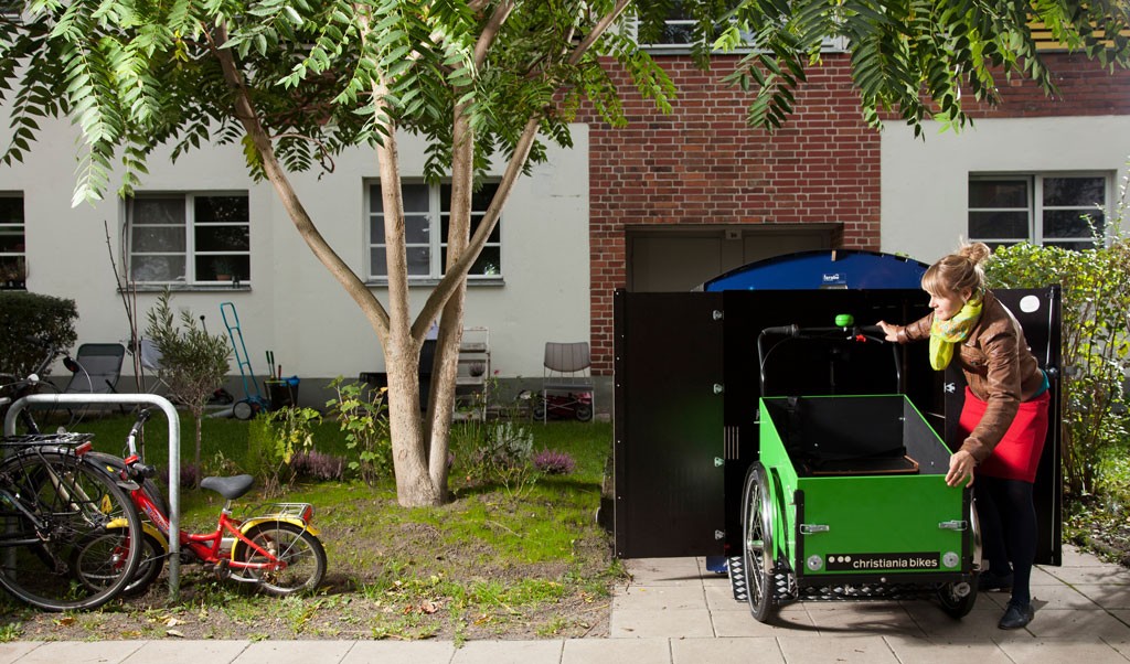 Frau mit grüner christiana Bikes Lastenrad vor schwarzem Abstellraum in urbanem Garten mit Baum und Fahrrädern