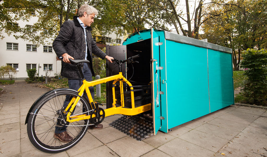 Mann lädt gelbes Lastenfahrrad in großen türkisfarbenen Fahrradcontainer an urbanem Standort
