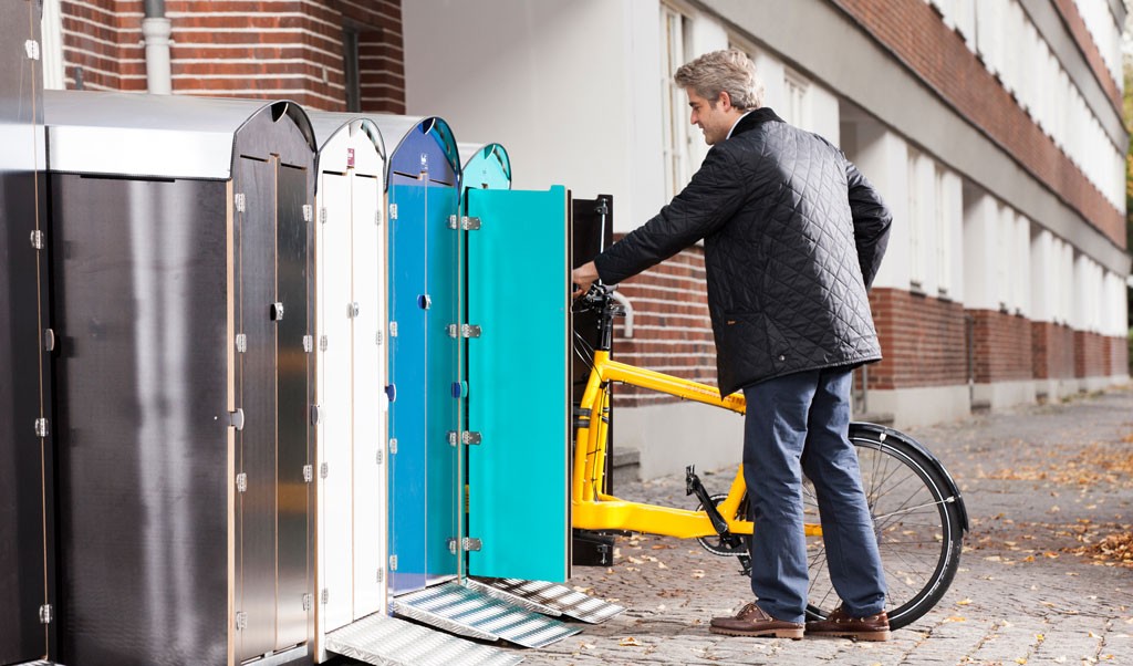 Mann stellt gelbes Fahrrad in farbige Abstellboxen aus Metall an urbanem Wohngebäude ab