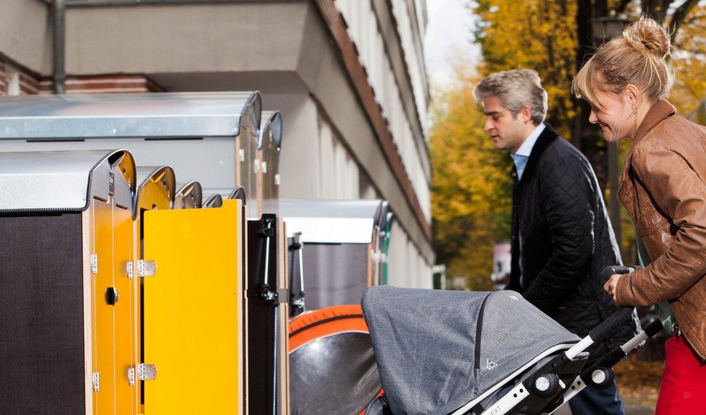 Paar mit Kinderwagen nutzt gelbe Kiwabo Paketboxen vor einem Wohnhaus im Herbst