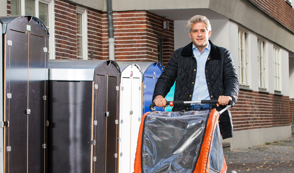 Mann mit schwarzer Steppjacke schiebt Kinderfahrradanhänger auf Gehweg vor Backsteinhaus