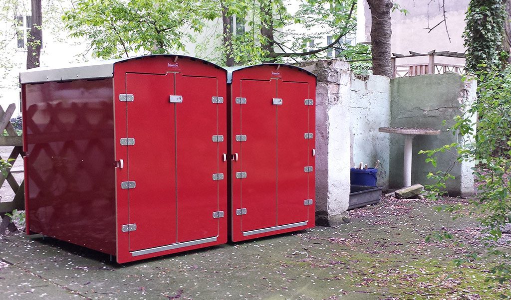 Zwei rote XXL Kiwi-Abfalltonnen nebeneinander in einem begrünten Außenbereich mit grünem Laub und altem Gemäuer