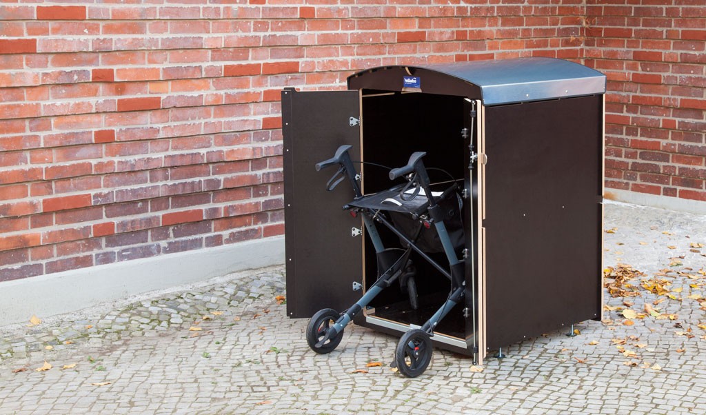 Schwarzer Rollator im geöffneten sicheren Außenbox aus Holz vor roter Backsteinwand