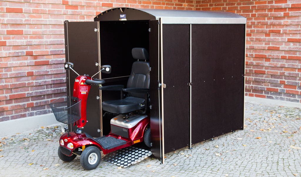 Elektromobil vor schwarzem Rollabo XXL Plus Unterstand mit offener Tür auf gepflastertem Boden vor roter Backsteinwand